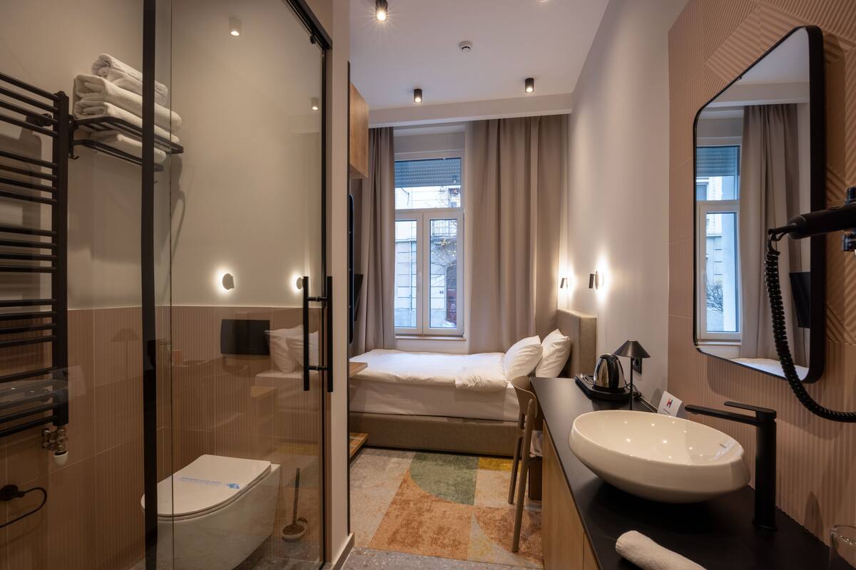A modern bathroom with a glass shower enclosure, a built-in toilet, a bedside table with a lamp, and a large mirror.
