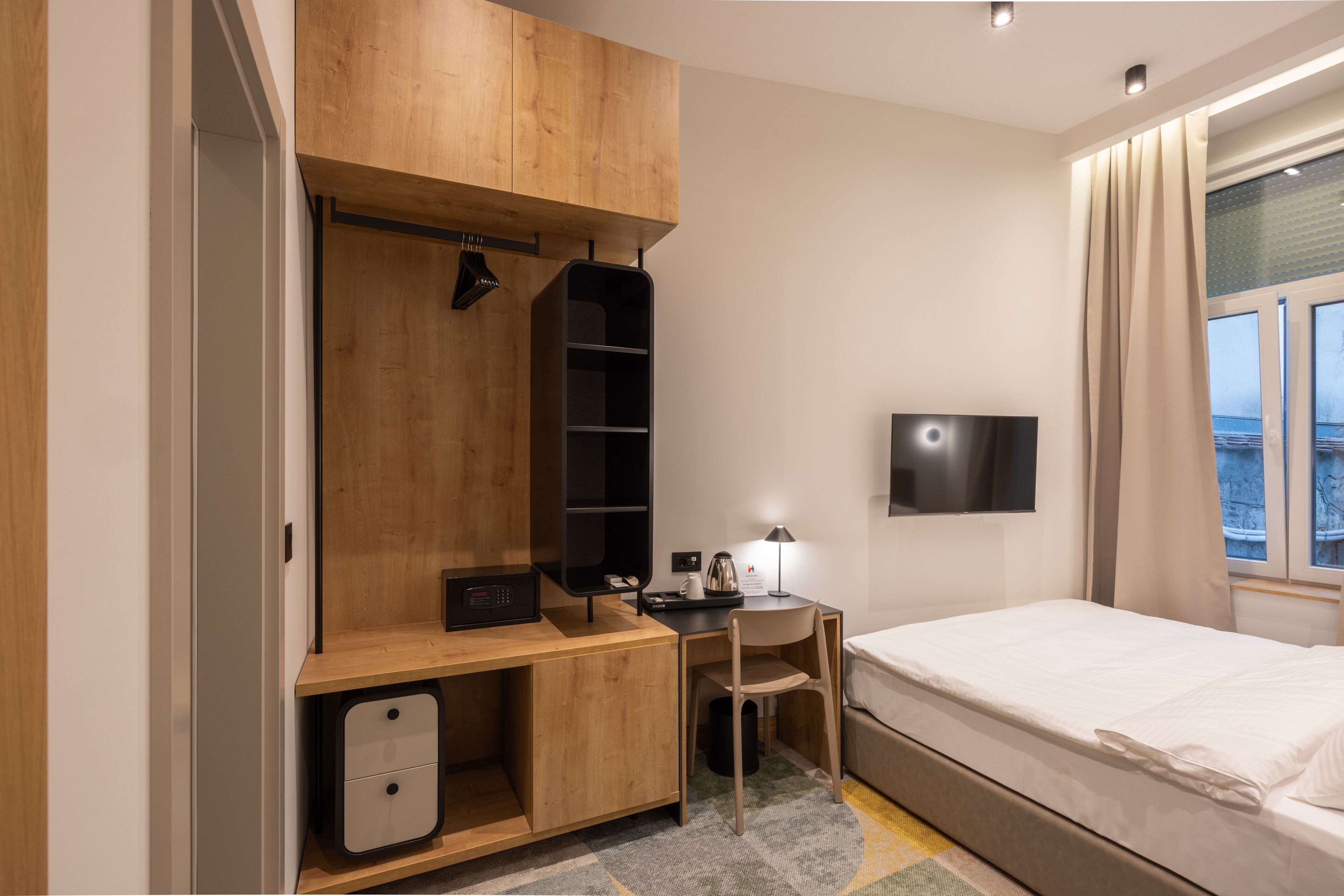 A modern hotel room with a wooden wardrobe, a desk with a lamp, a flat-screen TV, and a bed.