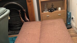 A room with a brown sofa bed, a blue rug, a wooden floor, a small wooden side table with decorative items, and a black chair.