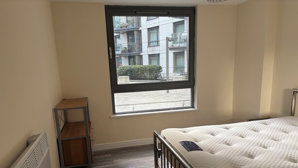 A bedroom with a single bed, a window, a radiator, and a wooden shelf.