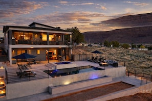 A modern two-story house with a large outdoor pool, outdoor seating, and a scenic mountain view. - Exclusive Retreats Desert Vista: Luxury 6 Bedroom Retreat- Heated Pool/ Hot tub (Moab)