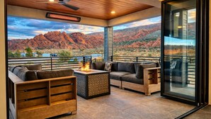 A modern outdoor seating area with a fire feature, wooden furniture, and a large glass door leading to a scenic view of red rock formations.