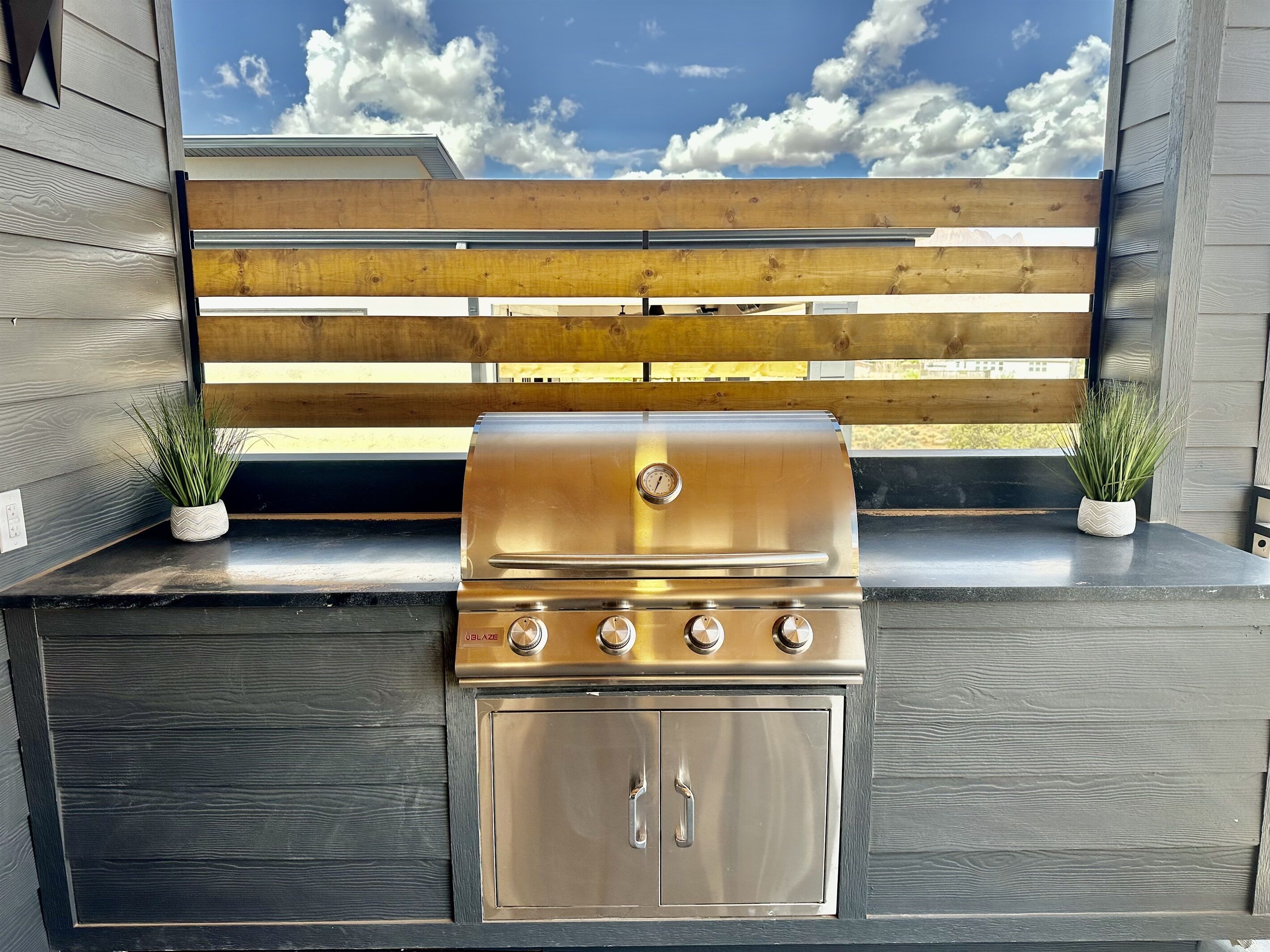 An outdoor kitchen with a stainless steel grill, wooden bench, and potted plants.