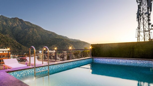 A swimming pool with a tiled edge, a metal bridge, and a mountain backdrop. - HOTEL VINSOBER PRIDE INN (Narendranagar)