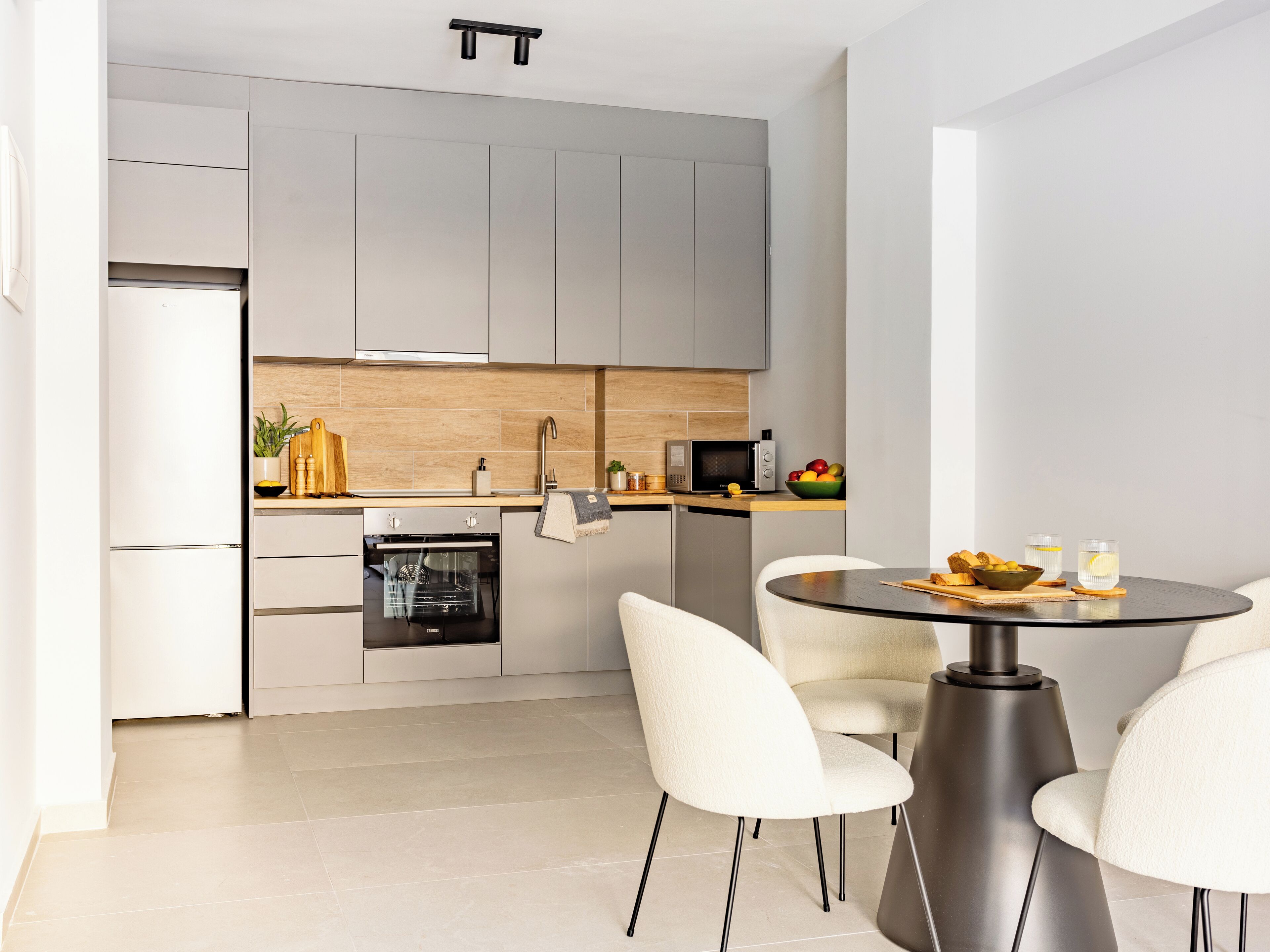Una cocina moderna con armarios grises, horno empotrado, microondas y una zona de comedor con mesa redonda y sillas blancas.