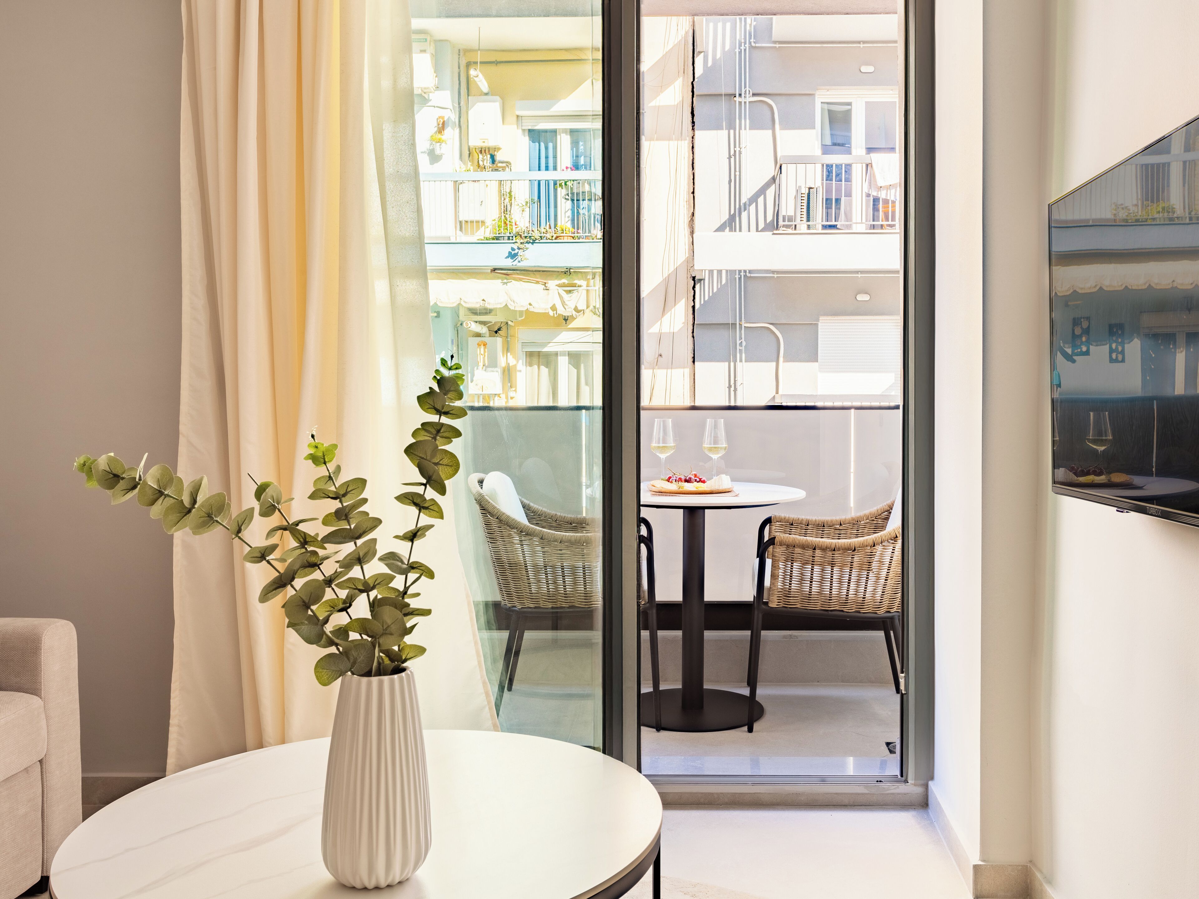 A modern room with a round table, a white vase with green foliage, and a view of a balcony with outdoor furniture and a dining table.