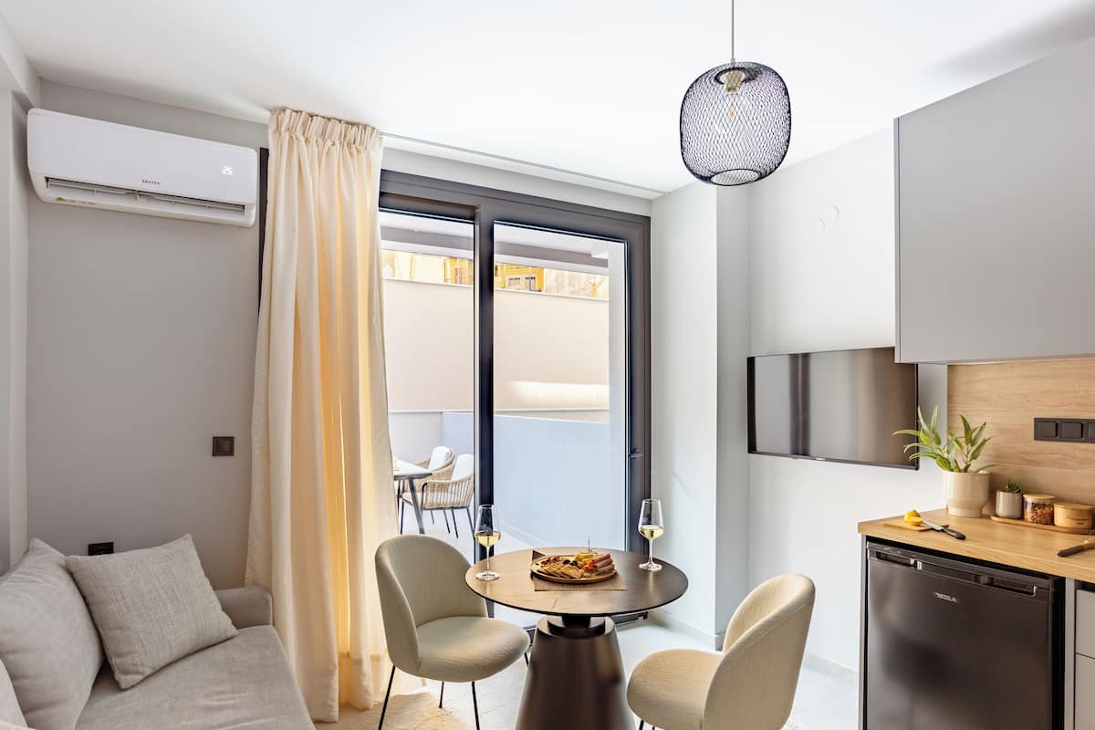 Una sala moderna con un sofá, dos sillones, una mesa de comedor con una copa de vino y una cocina con un refrigerador y una planta.