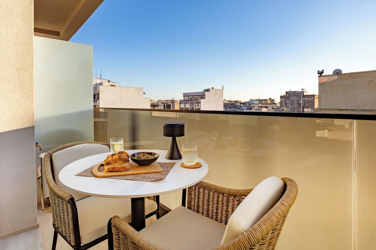 Una mesa redonda con mantel blanco, dos sillas de mimbre, un cuenco de aceitunas, un plato con pan y una vela en un balcón con vistas a la ciudad.