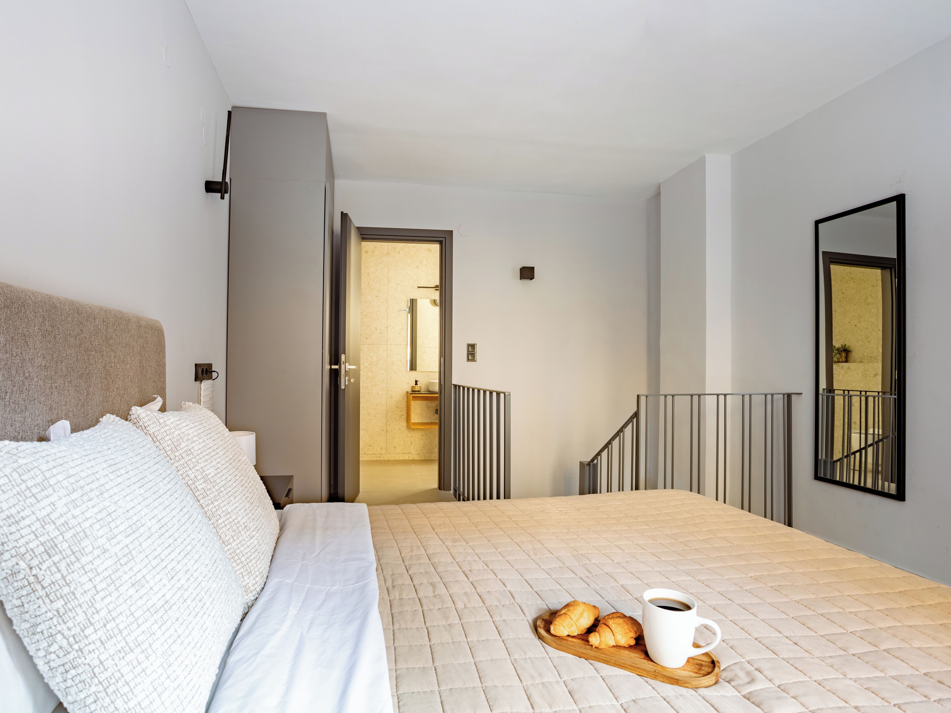 Une chambre avec un lit, un plateau contenant des croissants et une tasse de café, une salle de bain visible à travers une porte ouverte, et un escalier avec une rampe.