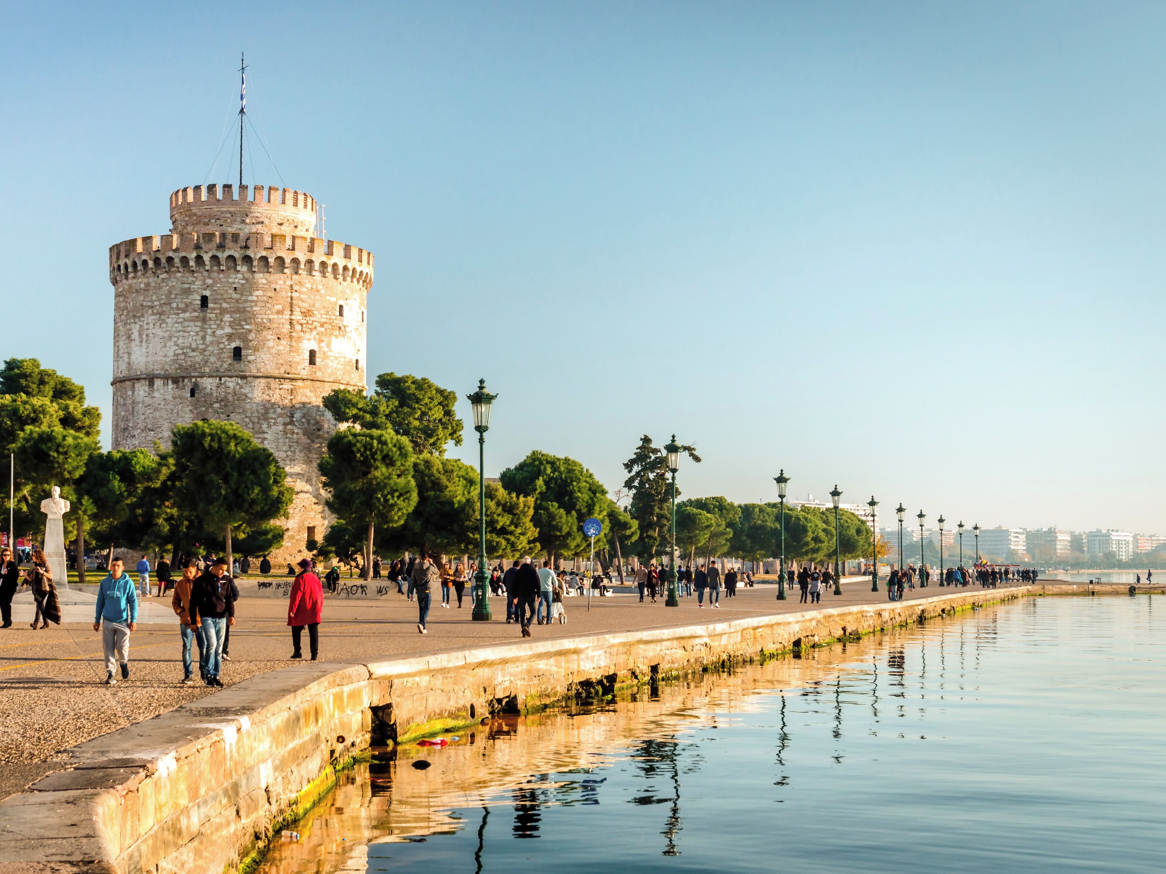 A historic tower with a circular base and a flat top, surrounded by a waterfront promenade with trees and lampposts.