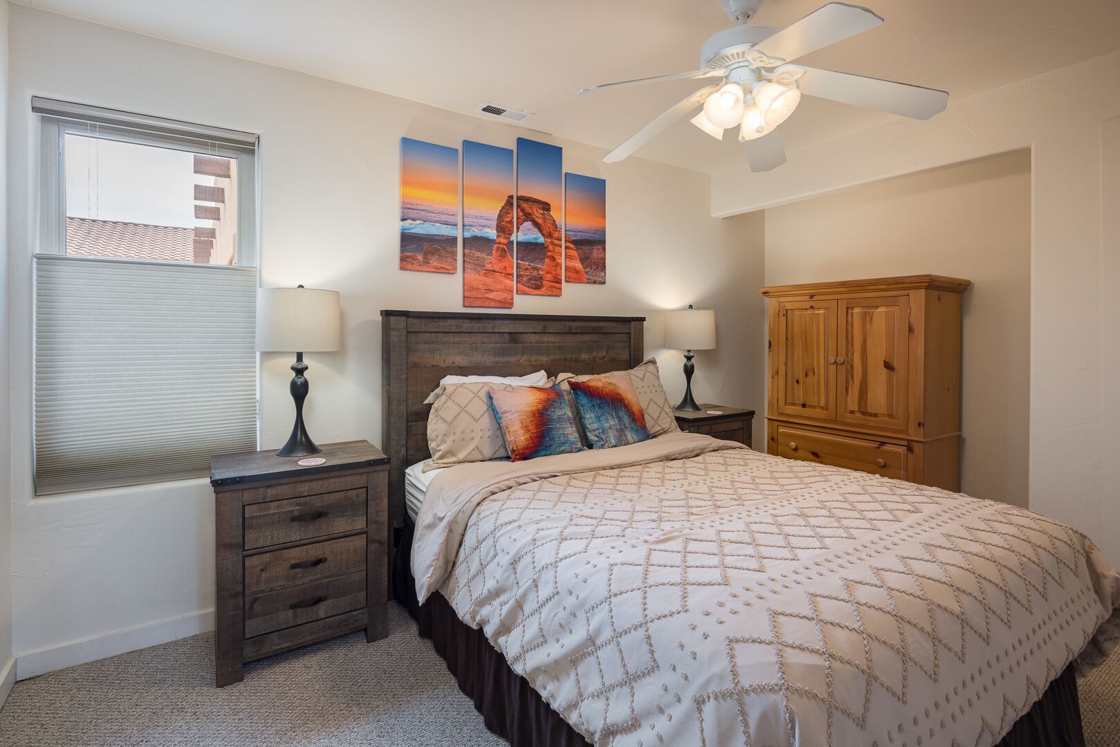 A bedroom with a bed, two bedside tables with lamps, a wooden wardrobe, a ceiling fan, and three sunset paintings on the wall.