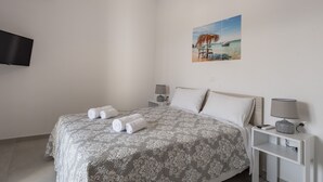 Un lit bien fait, recouvert d’une couvre-lit à motifs, avec deux serviettes enroulées, une table de chevet avec une lampe, un téléviseur fixé au mur et un tableau représentant une scène de plage.