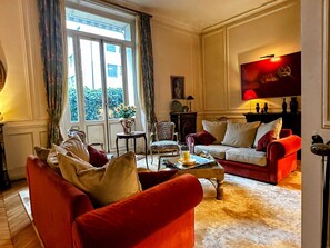 Ein Wohnzimmer mit einem roten Sofa, einem weißen Sofa und einem gemusterten Sessel. Ein Fenster mit Vorhängen, eine Vase mit Blumen und ein Bild an der Wand sind ebenfalls zu sehen.