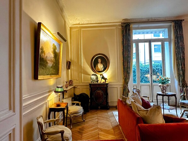 Une pièce avec un canapé rouge, un plancher en bois, une table avec une lampe et une fenêtre avec des rideaux.