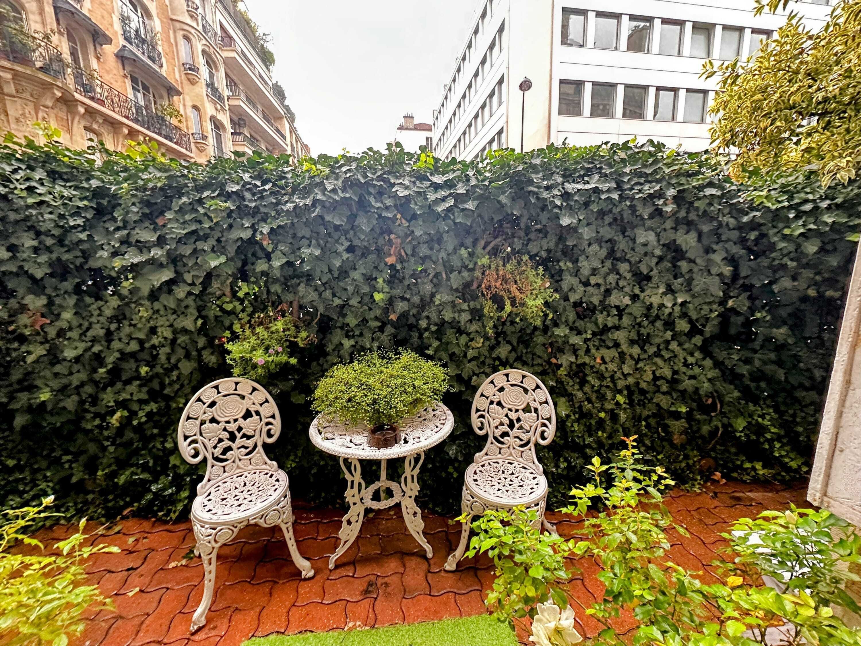 A garden area with a hedge, two ornate chairs, and a small table with a potted plant.
