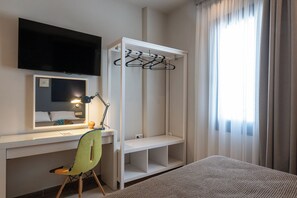 Une chambre d’hôtel équipée d’un bureau, d’une chaise, d’une télévision, d’une armoire et d’une fenêtre avec des rideaux.