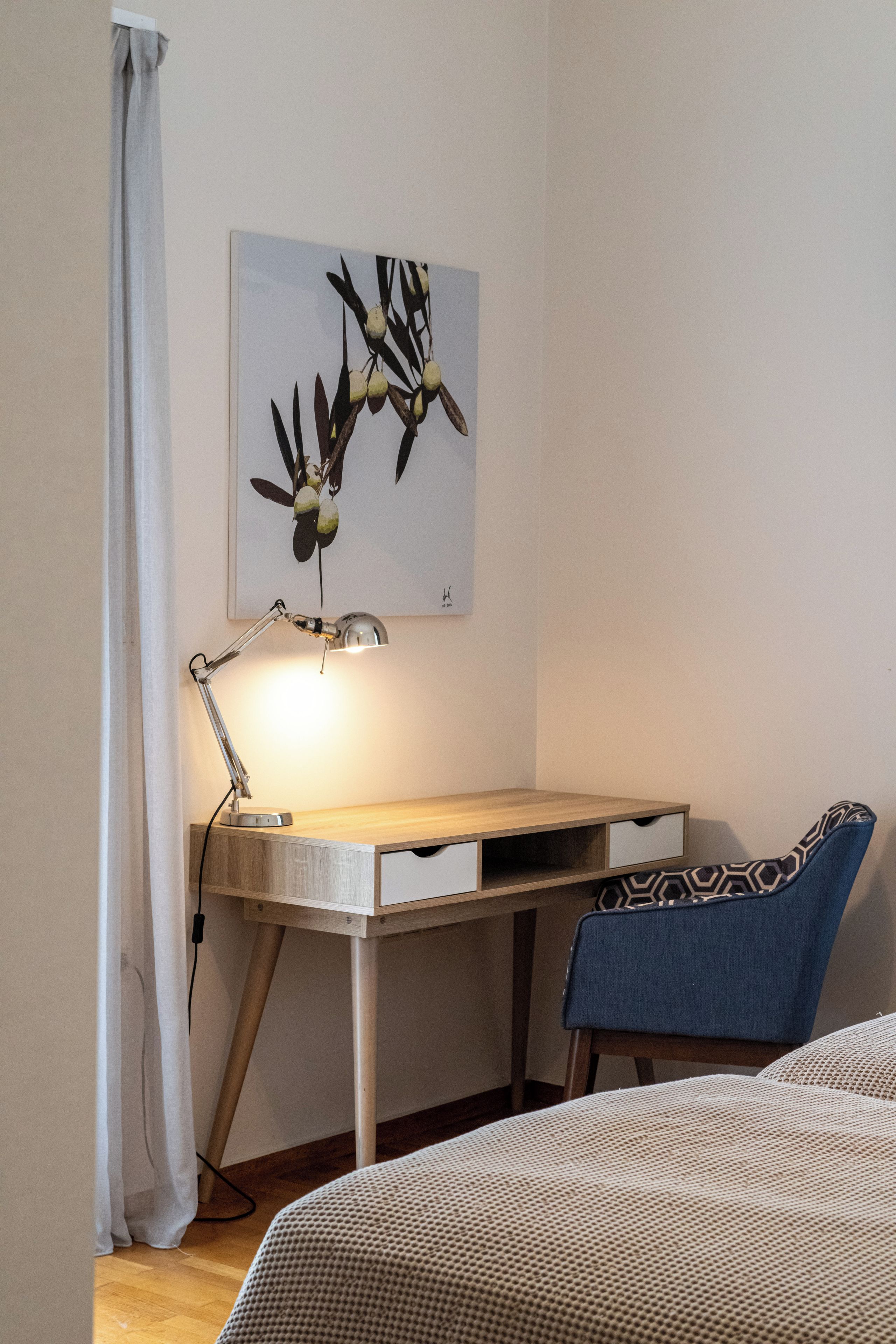 Un dormitorio con un escritorio de madera, una silla azul, una cama con cubrecama estampado, una lámpara de pared y una obra enmarcada sobre el escritorio.