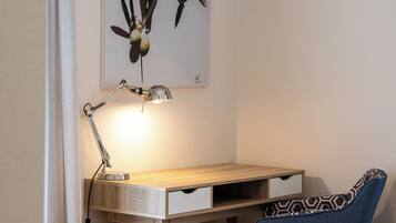 Un dormitorio con un escritorio de madera, una silla azul, una cama con cubrecama estampado, una lámpara de pared y una obra enmarcada sobre el escritorio.