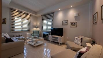 Una sala de estar moderna con sofá, sillón, mesa de centro, televisor, chimenea y ventanas grandes con persianas.