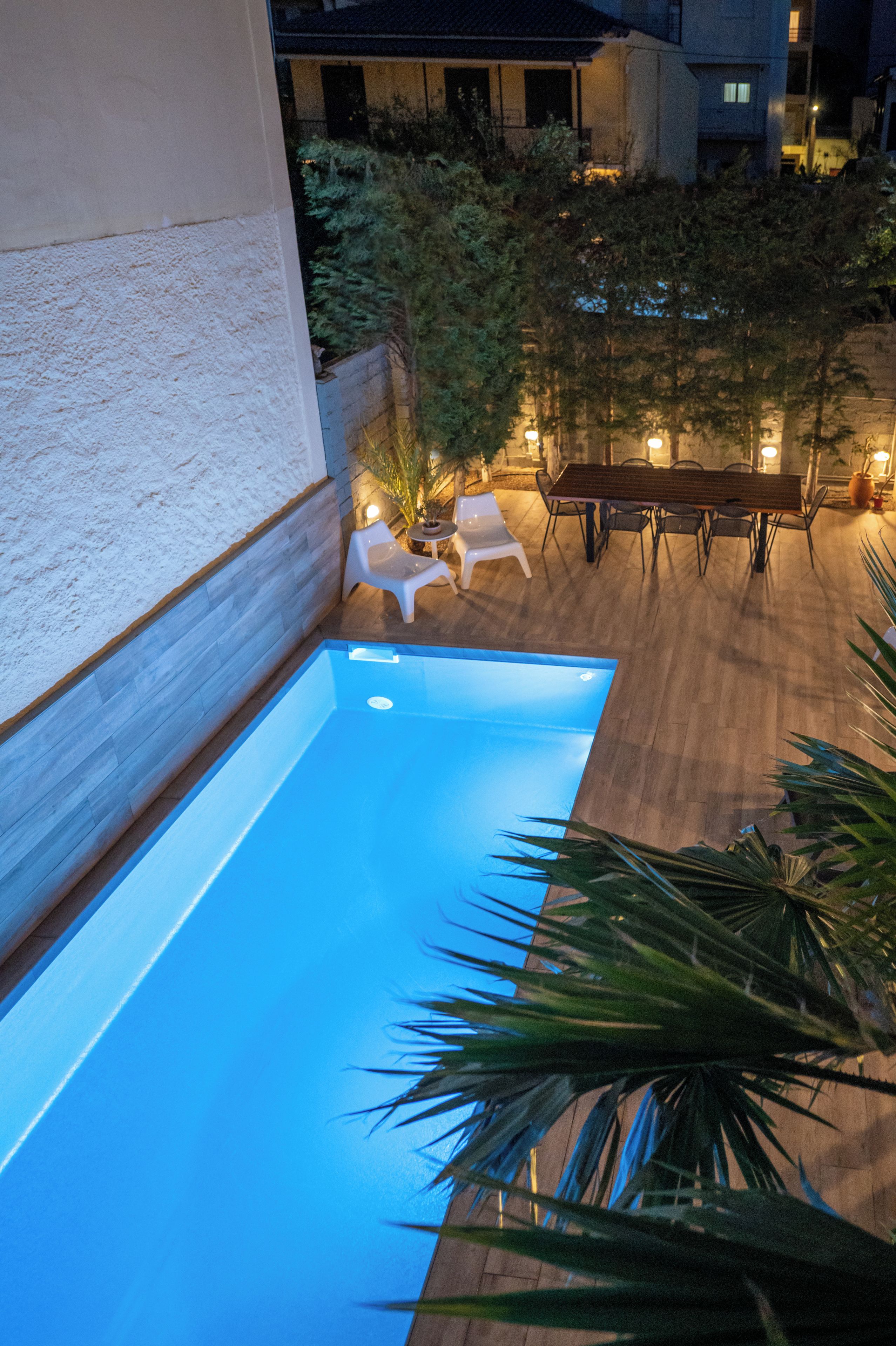 Una piscina esterna con bordo in legno, circondata da sedie e un tavolo, con un edificio sullo sfondo.