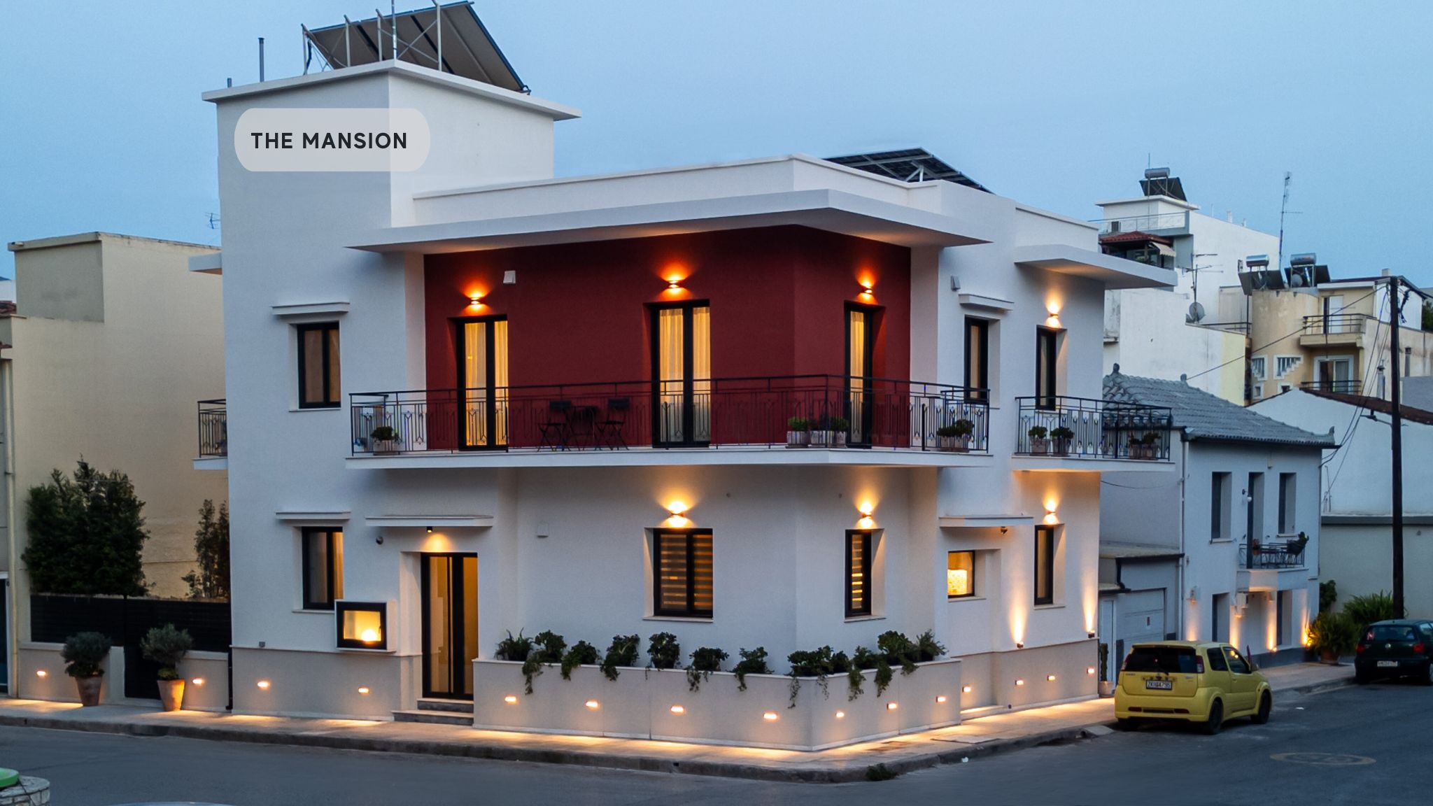 Um edifício moderno de dois andares chamado "The Mansion", com várias varandas, iluminação externa e um carro amarelo estacionado na frente.