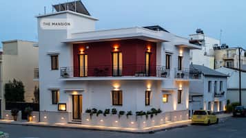 A modern two-story building named "The Mansion" with multiple balconies, outdoor lighting, and a yellow car parked in front.