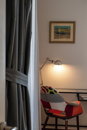 A chair with a colorful, patchwork design, a desk with a lamp, and a framed landscape painting on the wall.
