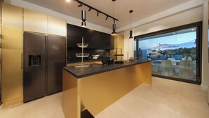 Une cuisine moderne dotée d’un îlot central, d’appareils électroménagers en acier inoxydable, de luminaires suspendus et offrant une vue sur l’extérieur grâce à une grande fenêtre.
