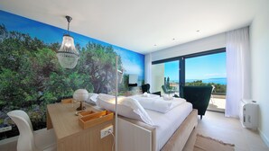Une chambre d’hôtel moderne avec un grand lit, un bureau agrémenté d’un globe terrestre et de quelques documents, une chaise, et une vue sur la mer par une grande fenêtre.