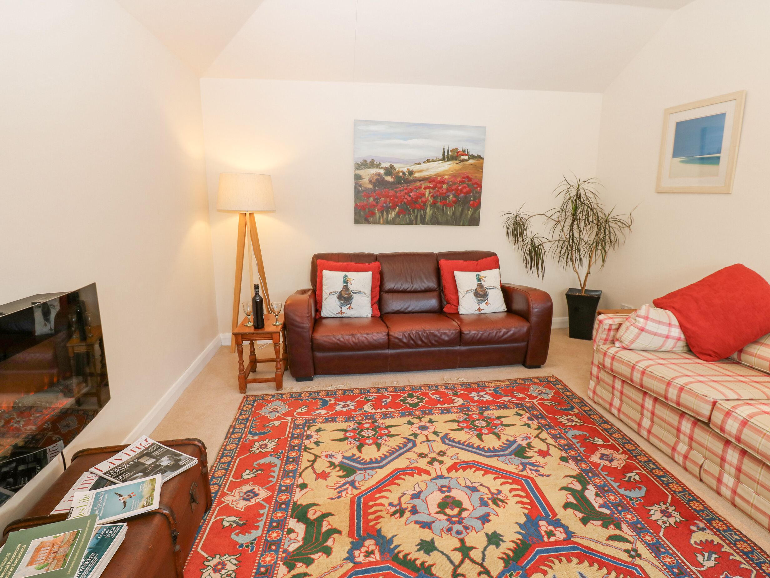 A living room with a brown leather sofa, a red and beige patterned rug, a wooden side table with a lamp, and a television on a stand.