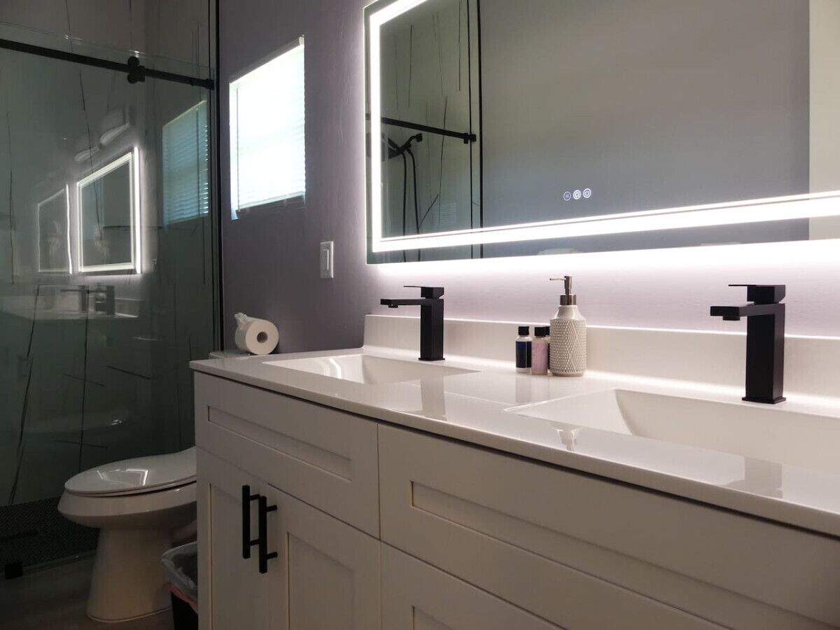 A modern bathroom with a white vanity, two black faucets, a large mirror with under-cabinet lighting, and a toilet.