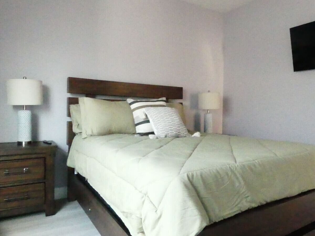 Een opgemaakt bed met een houten hoofdbord, een nachtkastje met een lamp en een wandlamp.