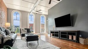 A spacious living room with a large TV mounted on the wall, a fireplace, and a view of the outside through arched windows. - Executive Suite (Marshall)