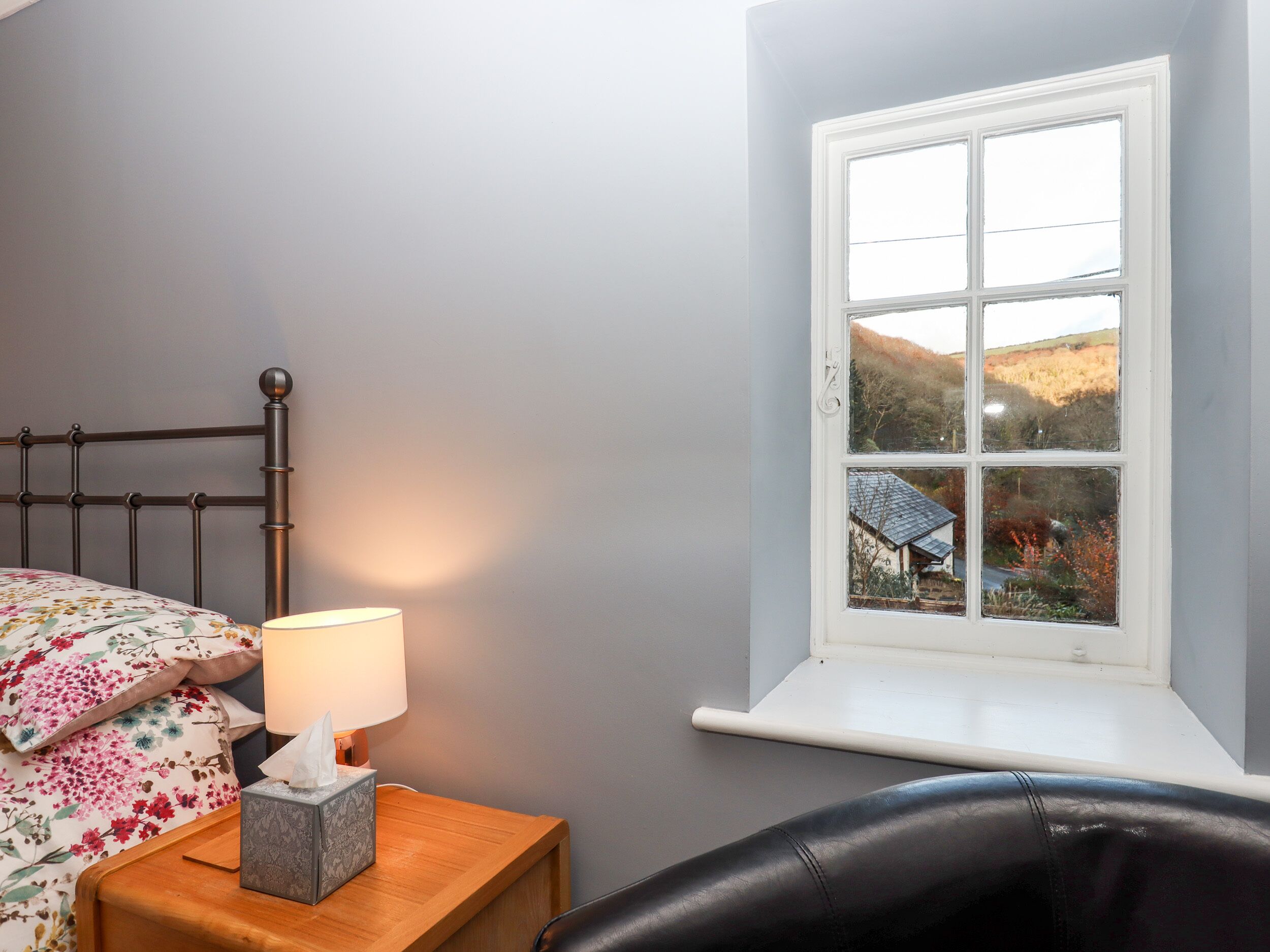 A bedroom with a bed, a nightstand with a lamp, and a window with a view of a house and trees.