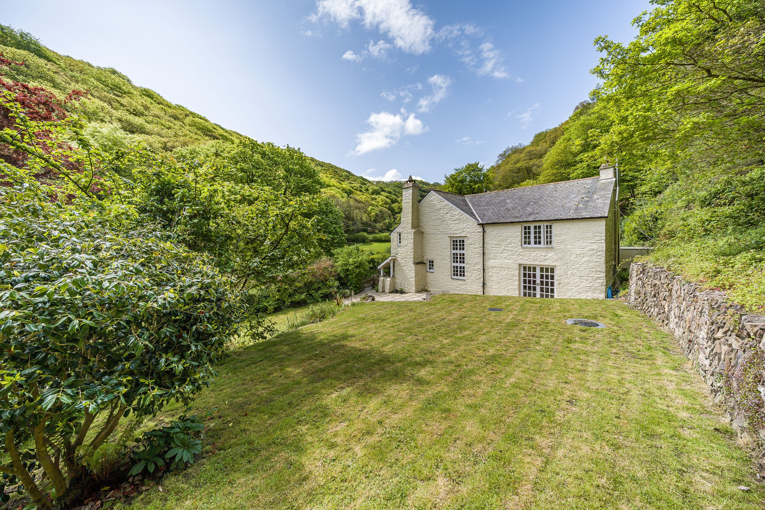 A stone house with a sloped roof, surrounded by lush greenery and a stone wall.
