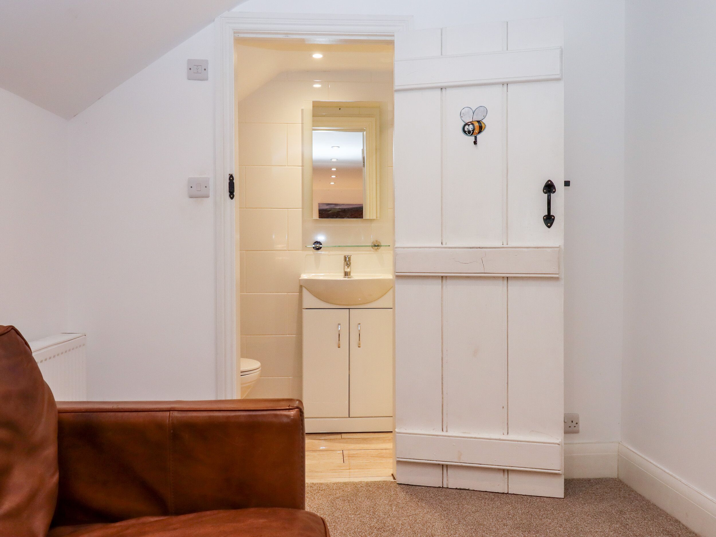 Una habitación con una butaca de cuero marrón, un armario blanco, un baño con lavamanos y espejo, y una puerta decorada con una abeja.