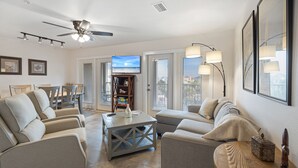 Ein geräumiges Wohnzimmer mit Deckenventilator, Fernseher und Blick auf den Strand durch die Schiebetüren.
