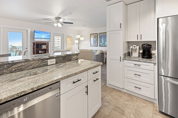A modern kitchen with granite countertops, white cabinetry, a stainless steel refrigerator, and a ceiling fan.