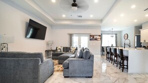 A spacious living area with a large gray sofa, a smaller gray sofa, and a coffee table. There is a kitchen area with a bar and stools, and a dining table with chairs.