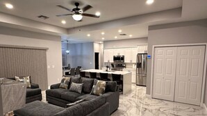 A modern living room with a grey sectional sofa, a dining area, and a kitchen with white cabinets and stainless steel appliances.