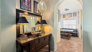 A hallway with a wooden console table, two black lamps, a mirror, and a chandelier. There is a bench with a patterned cushion and a dining area with a table and chairs in the background. - Elegant Beach Condo, Amazing Ocean Views, W/D (Galveston)