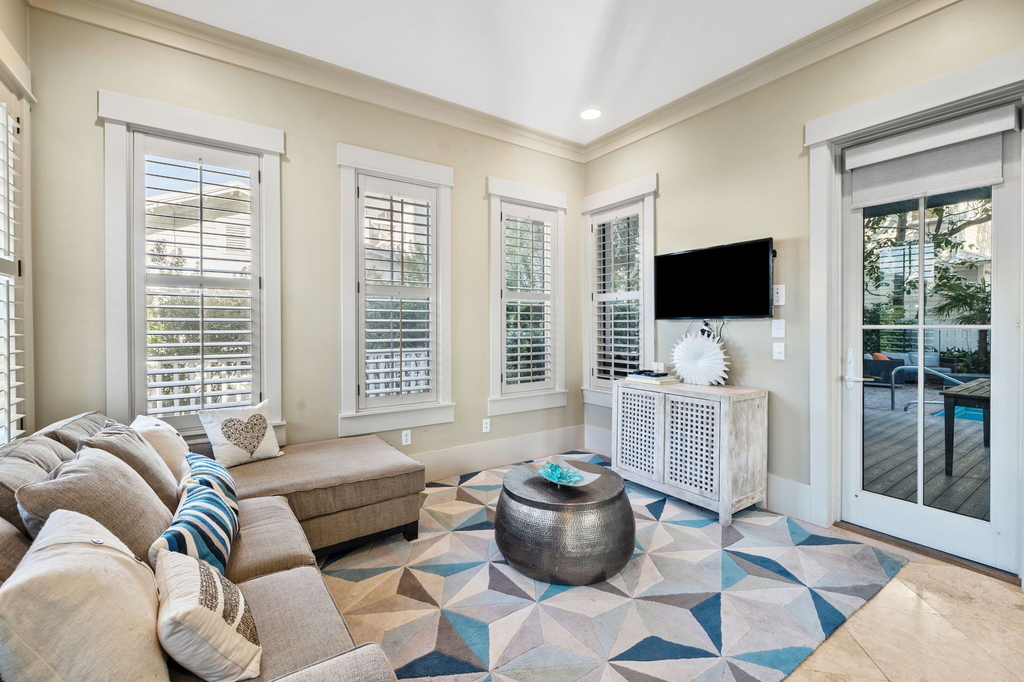 A sunlit living room with a beige sofa, a patterned rug, and a large window with blinds.