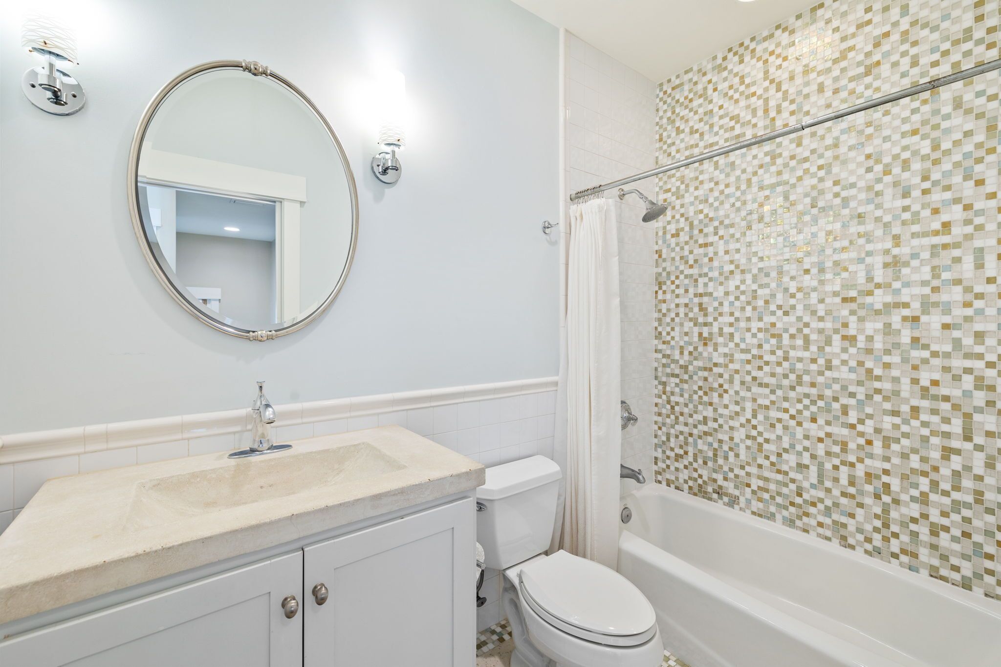 Une salle de bain avec un lavabo blanc, des toilettes, une douche dont le mur est recouvert de mosaïques et un miroir rond.