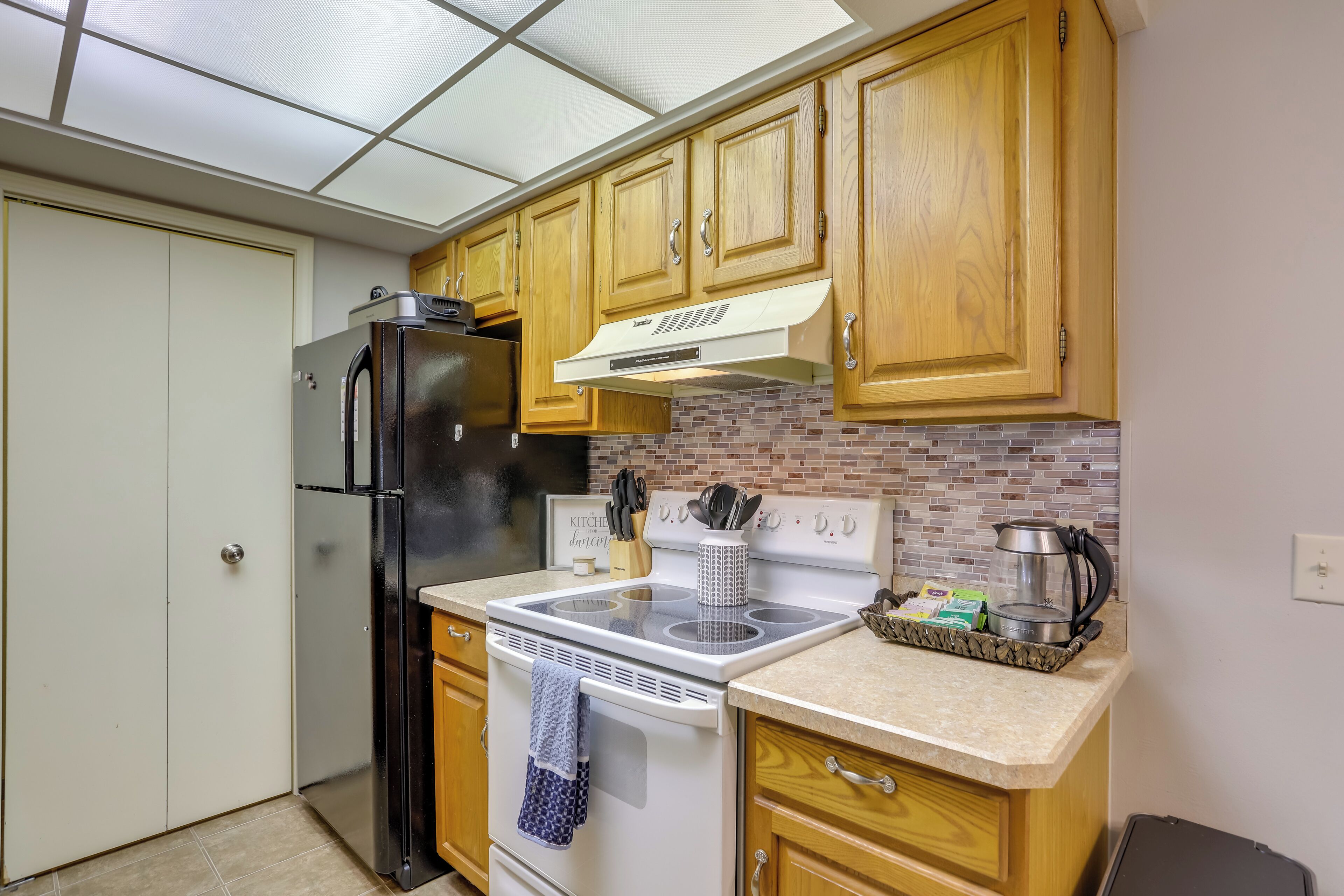 A compact kitchen with wooden cabinets, a black refrigerator, a white stove, and a brick backsplash.
