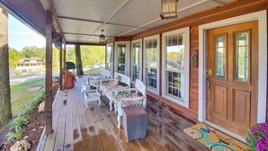A covered porch with wooden flooring, white and floral chairs, and a wooden door with glass panels.