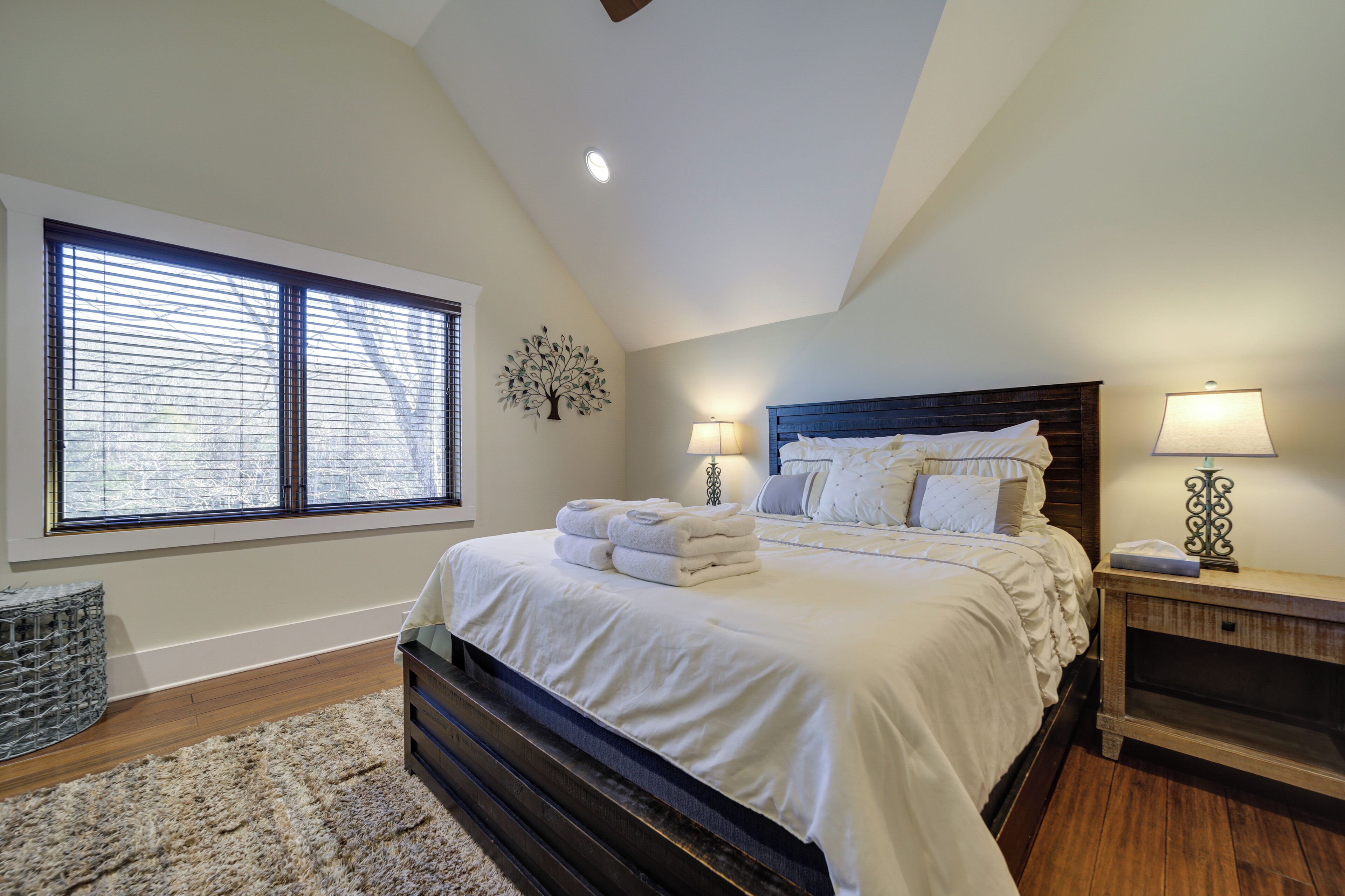 A bedroom with a large bed, two bedside lamps, a nightstand, a window with blinds, and a decorative wall piece.