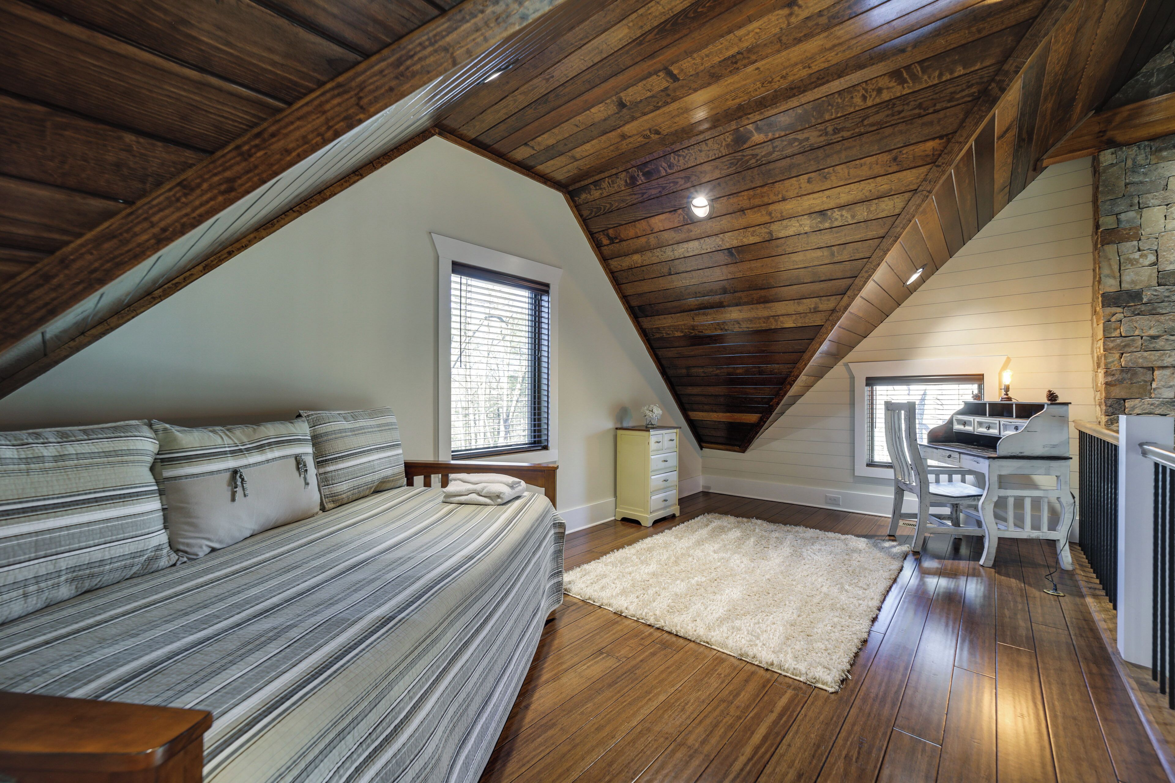 Un dormitorio con techo inclinado, una cama con ropa de cama a rayas, un escritorio con silla y una pared de piedra.