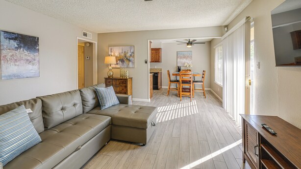 A living room with a sectional sofa, a wooden coffee table, and a ceiling fan. There is a dining area with a table and chairs, and a kitchen visible in the background. - Cozy Fresno Condo w/ Balcony & Pool Access (Fresno)