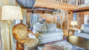 A spacious living room with a high ceiling, wooden beams, a white sofa, a wooden armchair, and a coffee table. - Charming Tamworth Cabin w/ Grill & Fireplace (Tamworth)