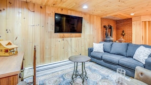 A cozy room with wooden walls, a blue sofa, a small round table, and a television mounted on the wall. - Charming Tamworth Cabin w/ Grill & Fireplace (Tamworth)
